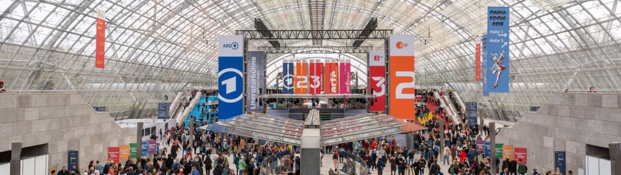 Innenaufnahme der Glashalle der Leipziger Messe mit bunten Werbebannern und vielen Besucher:innen während der Leipziger Buchmesse 2024